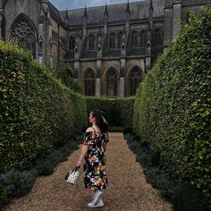 a woman in a floral dress is walking down a path between hedges and an old building