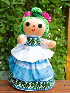 a doll with green hair and blue dress sitting on top of a wooden table in front of some bushes