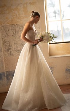 a woman in a wedding dress is standing by a window with flowers on her bouquet