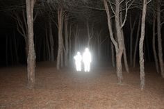 two people standing in the middle of a forest at night with light coming from their faces