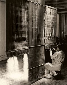 a woman kneeling on the floor in front of a large piece of art with fringes hanging from it