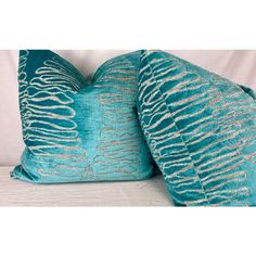 two teal and gold pillows sitting on top of a white couch next to each other