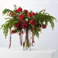 a vase filled with red roses and greenery on top of a white table next to a wall