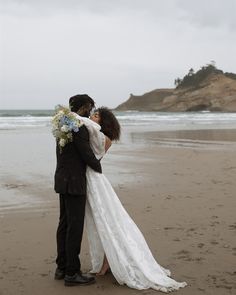 a bride and groom embracing on the beach in front of an ocean with cliffs behind them