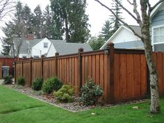 a wooden fence in front of a house next to a green yard with trees and bushes