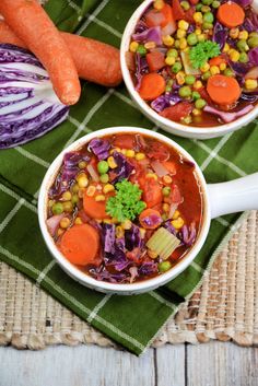 two white bowls filled with vegetable soup on top of a green cloth next to carrots and cabbage