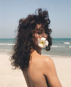 a woman with long curly hair and a flower in her mouth on the beach near the ocean