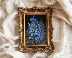 a painting with blue flowers in a gold frame on a white cloth covered wall behind it