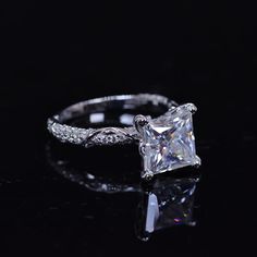 a princess cut diamond engagement ring on a black surface with reflection in the middle and an intricate filigree band