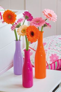 three different colored vases with flowers in them on a white table next to a bed