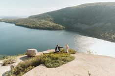 two people holding hands on top of a mountain overlooking a body of water and mountains