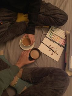 a person sitting on a bed holding a cup of coffee next to a notebook and pen