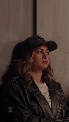 a woman with long hair wearing a black hat and leather jacket sitting against a wall