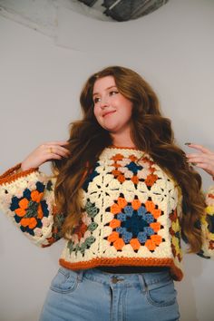 a woman with long brown hair wearing a crochet sweater and jeans standing in front of a white wall