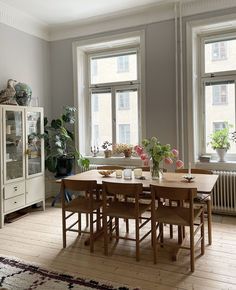 a dining room table and chairs in front of two windows with potted plants on them