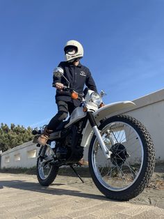 a man sitting on top of a motorcycle next to a white wall and trees in the background