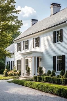 a large white house with black shutters on the front and side windows, surrounded by hedges