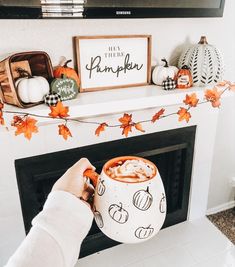 a person holding a mug in front of a fireplace with pumpkins on it and decorations