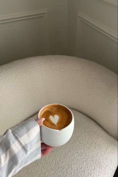 a person holding a cup of coffee on top of a white round couch with a heart drawn in the foam