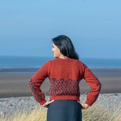 a woman standing in front of the ocean wearing a red sweater with an intricate design