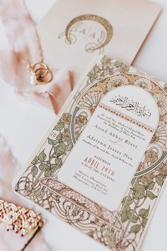 an ornate wedding card and ring on top of a pink satin ribbon next to it