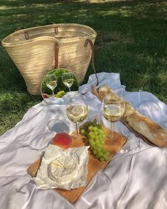 two glasses of wine, bread and grapes on a blanket in the grass near a basket