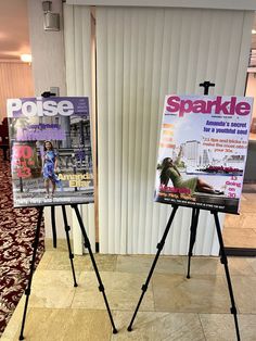 two posters are on easels in front of a wall with vertical blinds and a carpeted floor