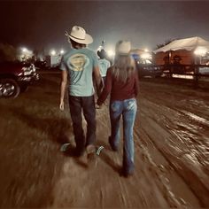 a man and woman walking down a dirt road at night with cars in the background