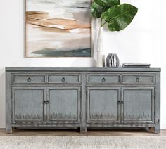 a large painting hangs on the wall above a gray buffet table with drawers and two vases