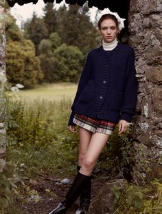 a woman standing in front of a stone wall with her legs crossed and boots on