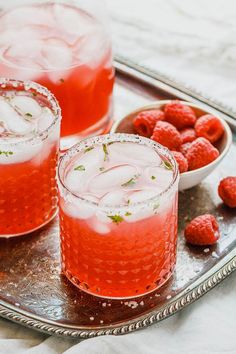 two glasses filled with raspberry lemonade on a silver tray next to bowls of raspberries