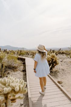 a woman in a white dress and hat walking across a wooden walkway with cacti