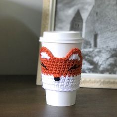 a coffee cup with a crocheted fox design on the front and side, sitting on a table next to a framed photograph