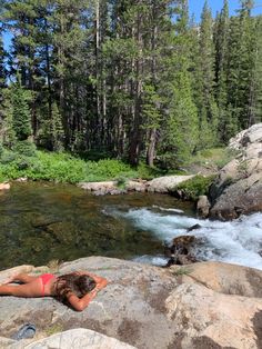 a woman laying on top of a rock next to a river surrounded by trees and rocks