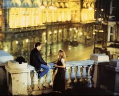 a man and woman sitting on a balcony overlooking a city at night with buildings lit up in the background