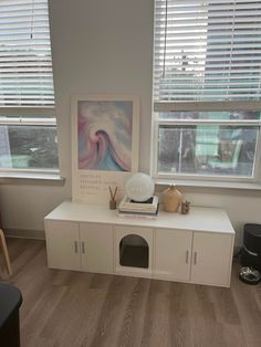 a room with two windows, a white cabinet and a painting on the window sill