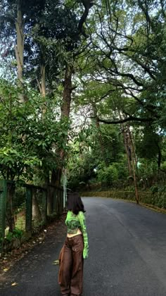 a woman standing on the side of a road in front of some trees and bushes