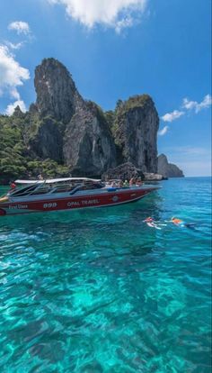 a red and white boat in clear blue water with people swimming near the rock formations