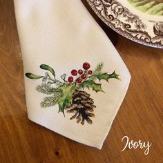 a white napkin with holly and pine cones on it sitting next to a dish towel