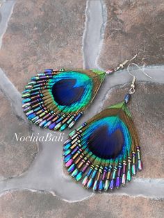 a pair of peacock feather earrings with multicolored beads on them sitting on top of a stone slab