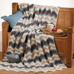 a crocheted blanket is sitting on a wooden bench next to a basket with food