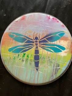 a colorful dragonfly painted on top of a round metal plate with trees in the background