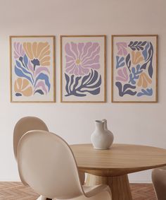 three paintings hang on the wall above a round table with two chairs and a vase