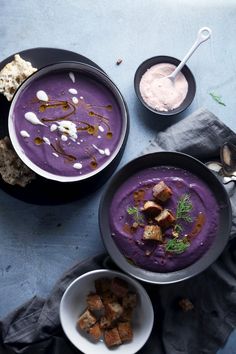 two bowls filled with purple soup next to bread and spoons on a blue surface