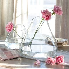 three pink roses are in a clear vase on a table with plates and napkins