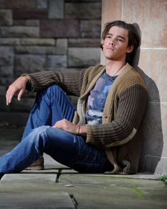 a young man sitting on the ground next to a brick wall with his hands in his pockets