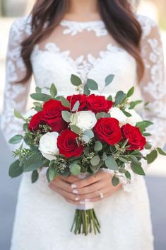 a bride holding a bouquet of red and white roses in her hands while wearing a lace wedding dress