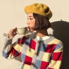 a woman wearing a yellow hat drinking out of a cup while standing next to a wall