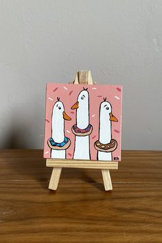 an easel with a painting of two ducks on it's sides and one duck in the middle holding a donut