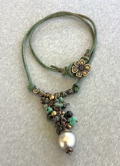 A shimmering almond glass pearl (12mm) hangs underneath green turquoise nuggets together with tigers eye and petrotourmaline rounds and brass beads. This complex hangs off green leather, together with brass beads either side. The necklace closes with a brass flower button and measures 17.5". Bohemian Green Jewelry With Pearl Charm, Adjustable Green Turquoise Pendant Necklace, Green Bohemian Necklace With Pearl Charm, Bohemian Green Necklace With Pearl Charm, Leather Cord Jewelry, Leather And Brass, Flower Button, Cord Jewelry, Brass Beads